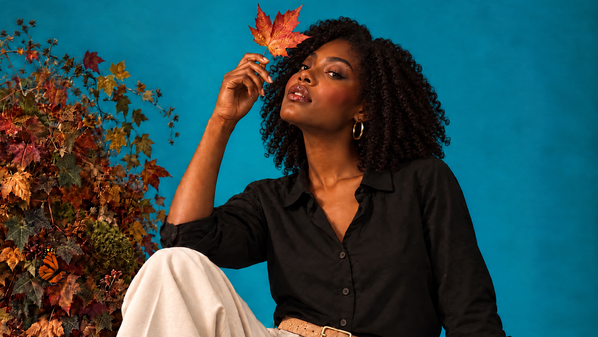 A woman with curly black hair sits on a blue studio floor surrounded by autumn leaves and greenery, wearing a black button-up shirt, cream wide-leg pants, and a tan belt. She holds a red maple leaf near her face against a bright blue backdrop.